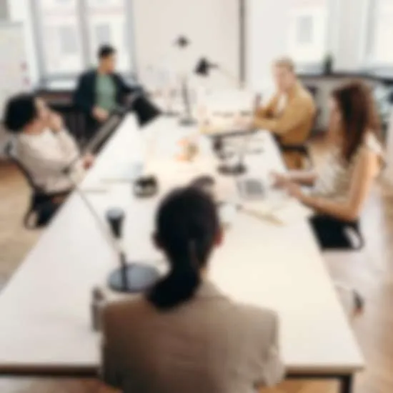 A blurred photo of a leadership team of five people sitting around a large, white office table in a modern workspace with a collaborative atmosphere. Laptops, papers, and coffee cups are spread out on the table. 