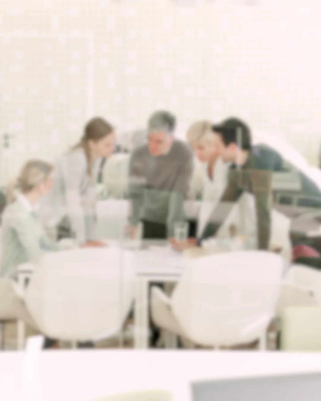 In a modern, light-toned office, a focused group gathers around a table, engaged in strategy and leadership development discussions. Leaning forward over documents and a laptop, they sit on white chairs, embodying effective business consulting services.