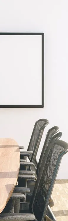 A conference room with a wooden table surrounded by black mesh chairs. A large blank whiteboard is mounted on the white wall in the background. Sunlight casts shadows across the table and floor.