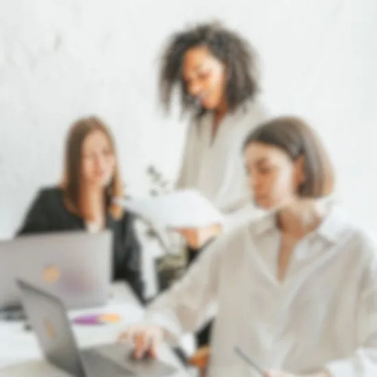 Three women are engaged in a team meeting at a table with laptops open. One person is standing, holding papers, while the other two focus on their screens. 