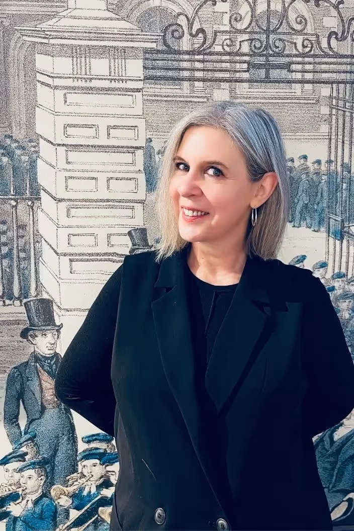 Catrina Hewitson, wearing a black outfit, smiles while standing in front of a vintage illustration featuring people in old-fashioned attire and top hats, embodying the timeless elegance and tailored strategies that define effective leadership.