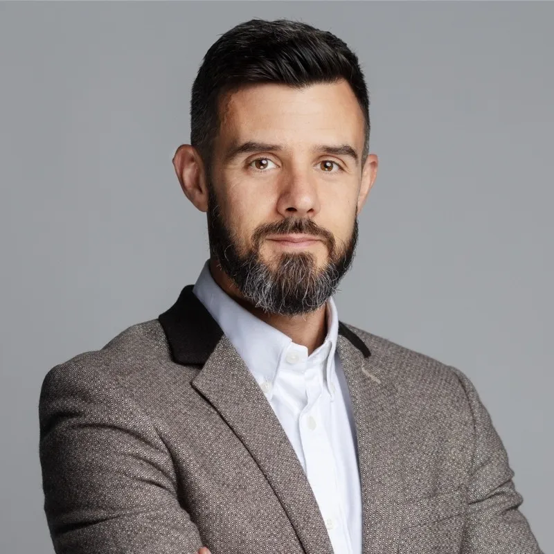 Andy Taylor stands confidently in front of a grey background wearing a grey suit and white shirt
