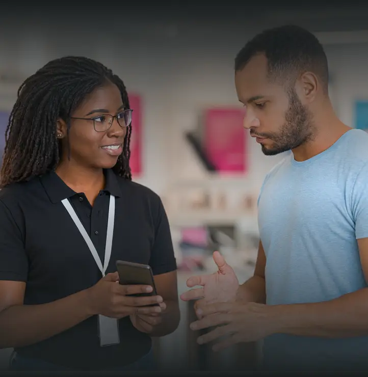 A woman in a black polo shirt with a lanyard shows a smartphone to a man in a light blue shirt inside a store.