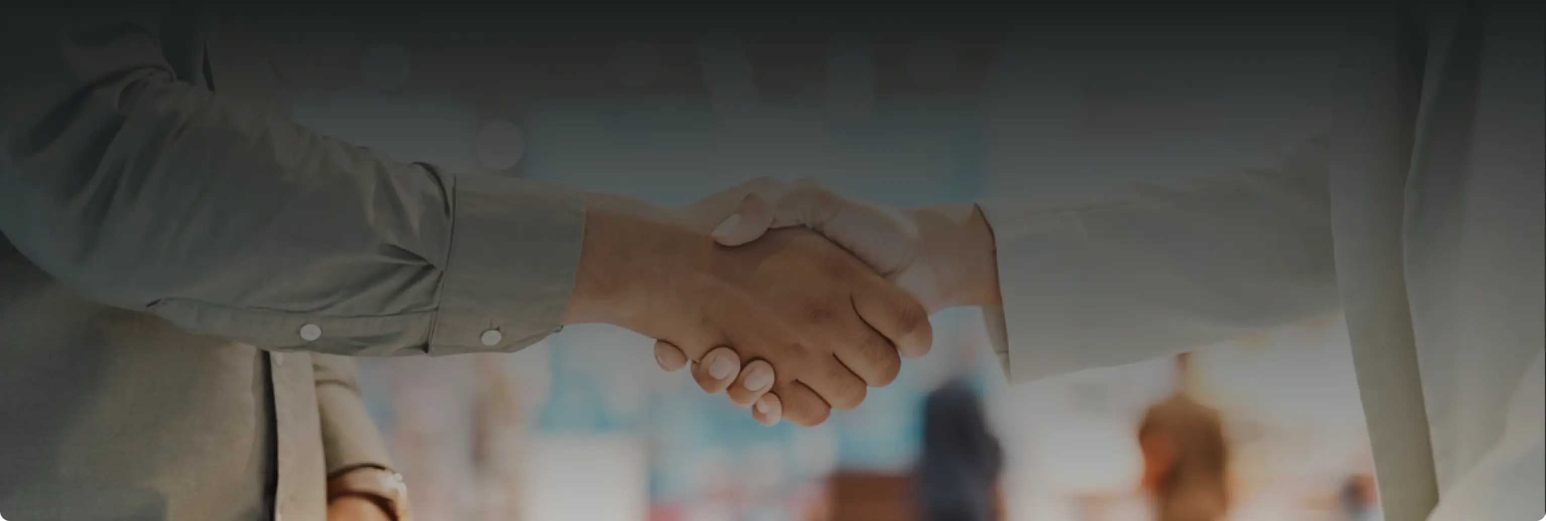 Two people shaking hands in a professional setting with blurred background figures.