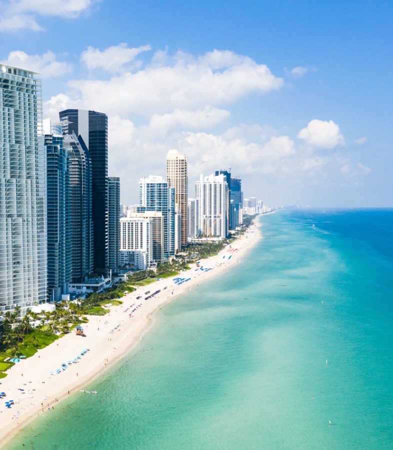 Aerial view of a sandy beach with turquoise water and tall city skyscrapers under a blue sky.