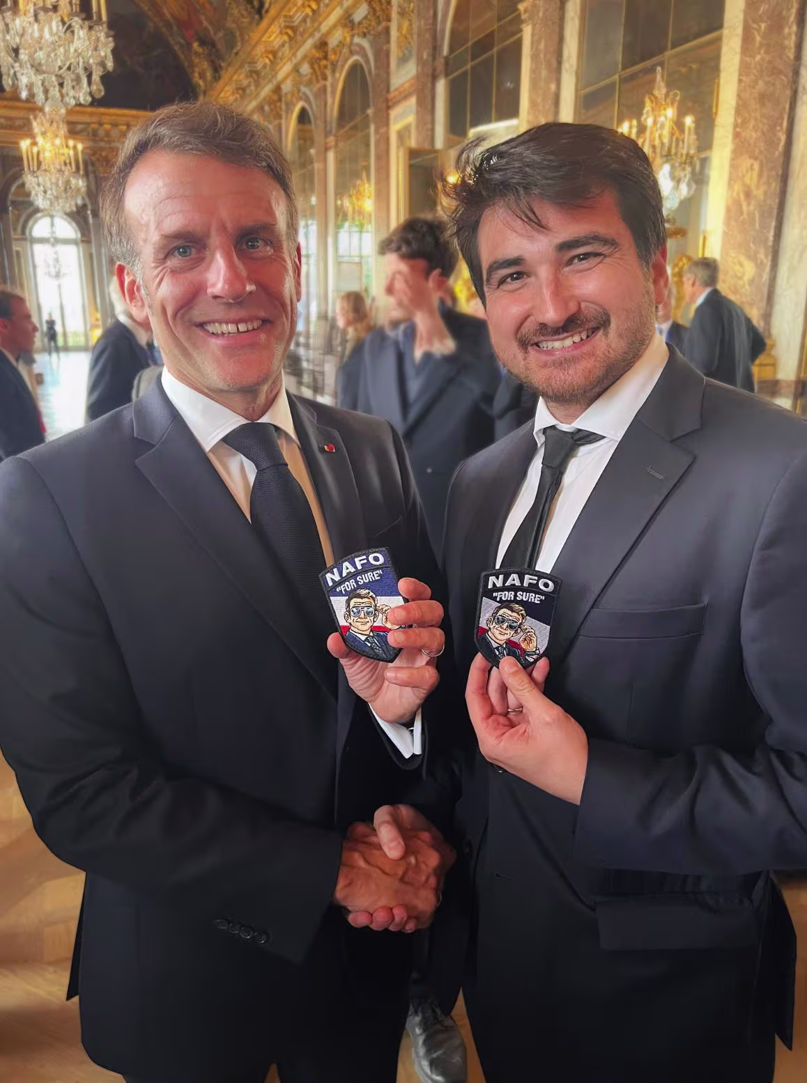 Two men in suits smiling and holding matching NAFO "For Sure" patches.