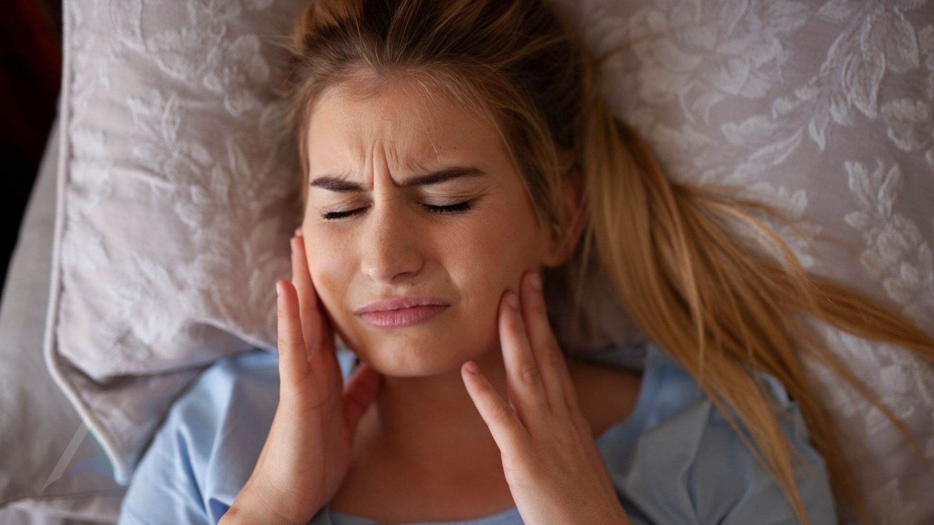 Person pressing a hand against the jaw in front of the ear, illustrating TMJ pain and its link to the upper neck