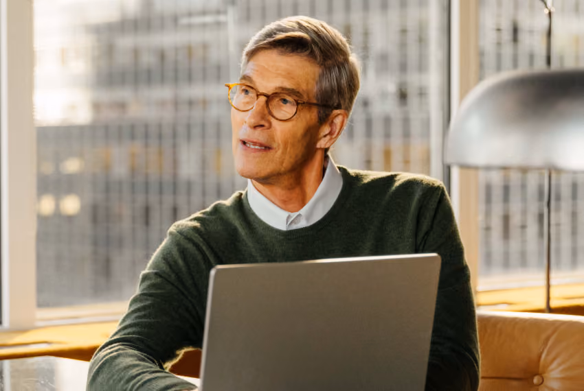 Middle-aged man with glasses and gray hair wearing a green sweater, sitting indoors in front of a laptop and looking thoughtfully to the side.