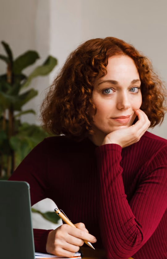 Woman with curly red hair wearing a burgundy sweater, sitting at a desk with a laptop, holding a pen and resting her chin on her hand, with green plants in the background.