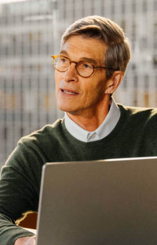 Middle-aged man with glasses wearing a green sweater over a white collared shirt, sitting indoors with a laptop in front of him, looking to the side thoughtfully.