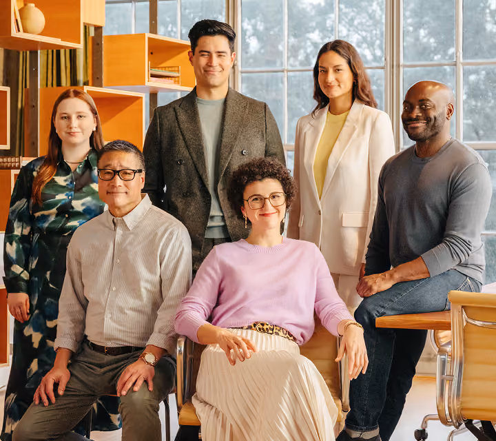 Portrait of six diverse professionals in a modern office, with three standing behind and three seated, all smiling at the camera.