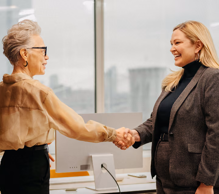 Two professional women smiling and shaking hands in a modern office setting with a computer and large windows in the background.