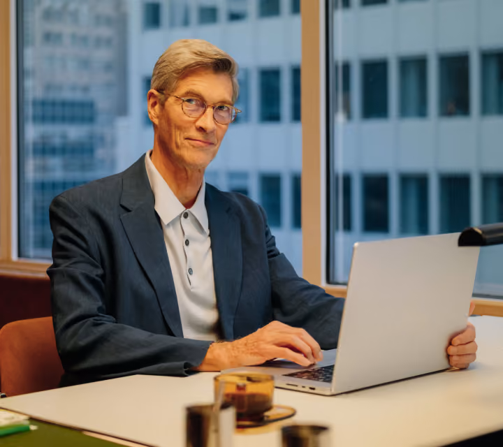 An older man with gray hair and glasses wearing a dark blazer and light shirt sits at a table in an office with large windows, typing on a laptop and looking at the camera.