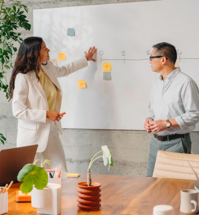A woman in a white suit explains a timeline drawn on a whiteboard to a man wearing glasses and a light shirt in a modern office setting with plants and a wooden table.