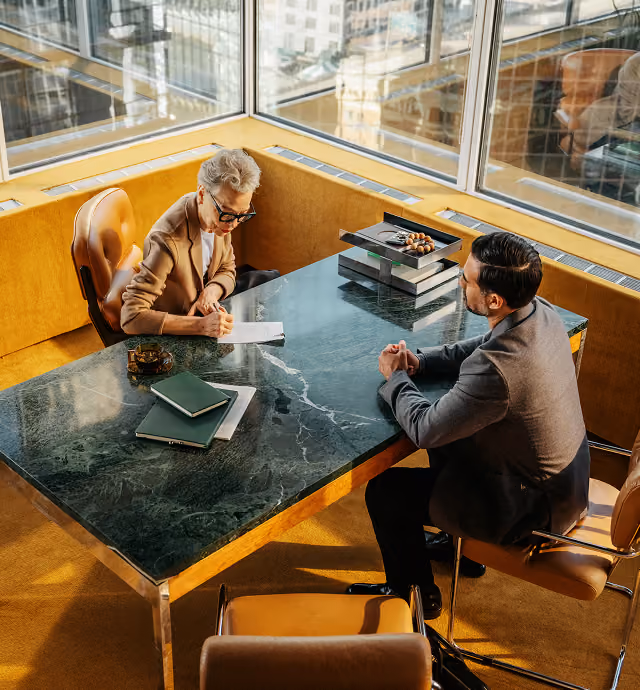 Two professionals in a modern office with large windows; a woman with short gray hair and glasses sits at a marble table writing on a notepad, while a man in a suit sits opposite her with hands clasped, engaging in discussion.