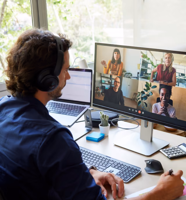 A man wearing headphones participates in a video conference on a desktop monitor showing four colleagues in separate windows, sitting at a desk with a laptop, keyboard, mouse, calculator, and small potted cactus nearby.