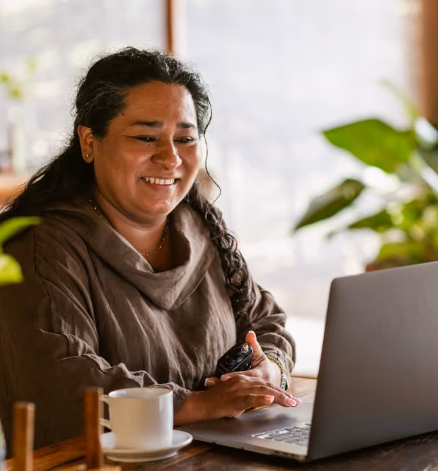 Smiling woman with dark hair working on a laptop at a wooden table with a white coffee cup nearby in a cozy indoor space with plants in the background.