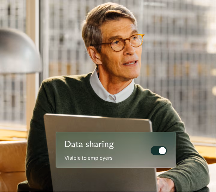Older man with gray hair and glasses wearing a green sweater sitting indoors near a window, looking to the side, with a laptop in front showing a 'Data sharing' toggle switch turned on and labeled 'Visible to employers'.