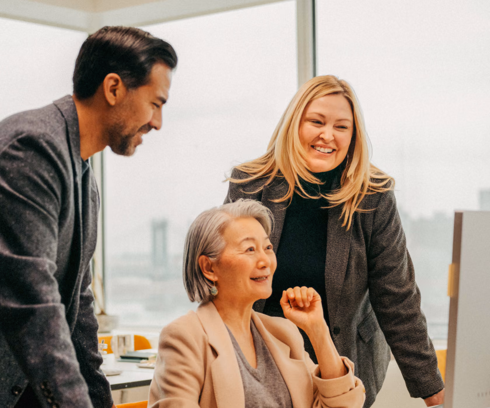 Three diverse business professionals smiling and engaging together in an office with large windows overlooking a city view, gathered around a desk with a computer and papers.