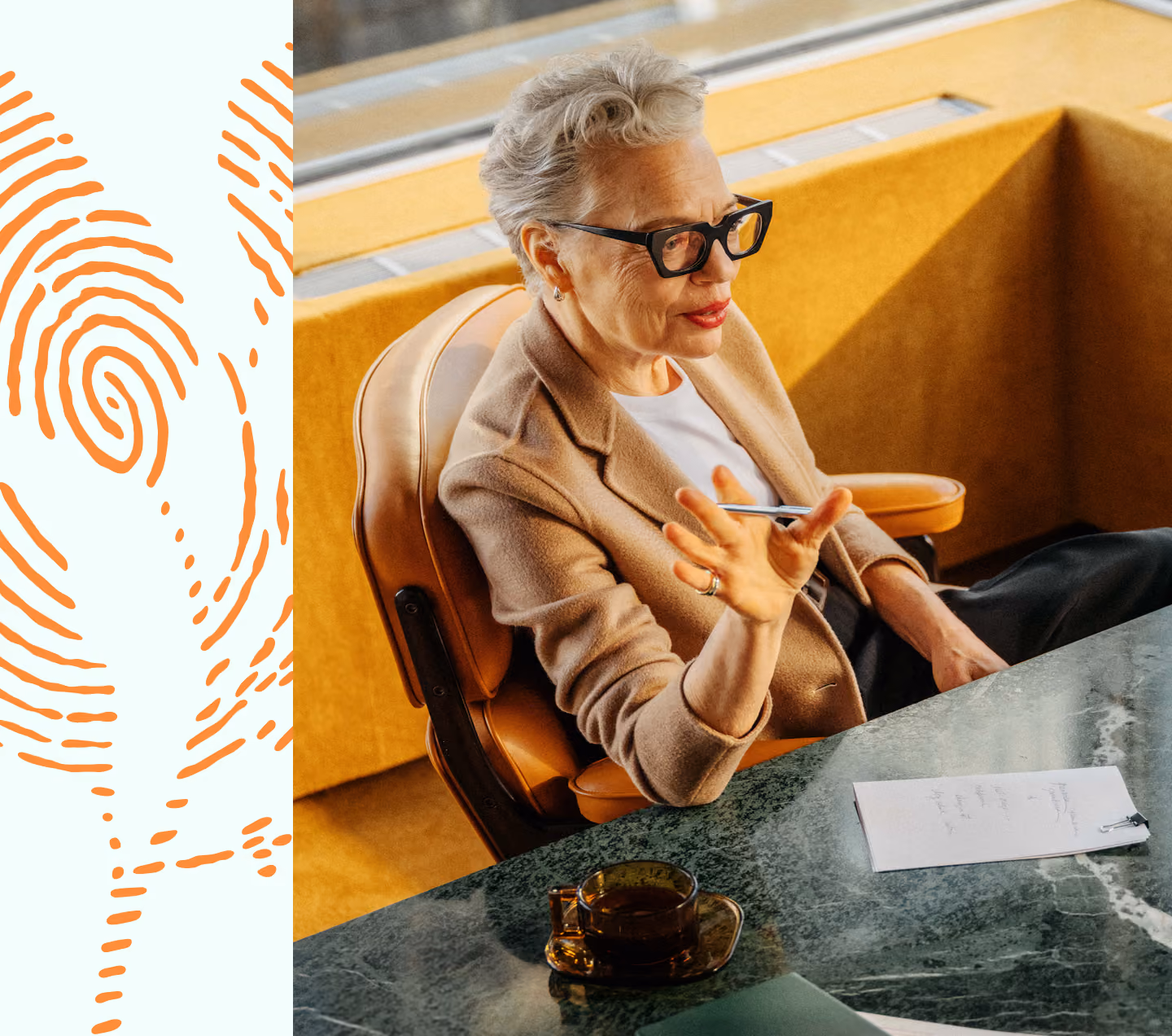 An older woman with short gray hair and black glasses sits at a marble desk holding a pen, gesturing with her hand. She wears a beige blazer over a white shirt. On the desk are a cup of tea and a notepad with writing.