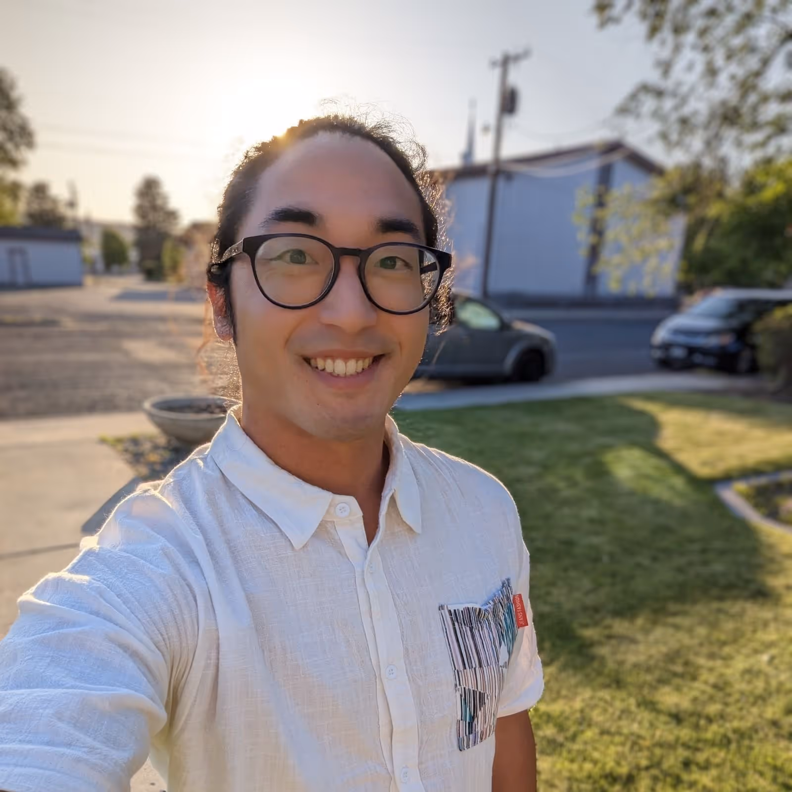 Smiling man with glasses wearing a white shirt standing outside in a sunlit yard.