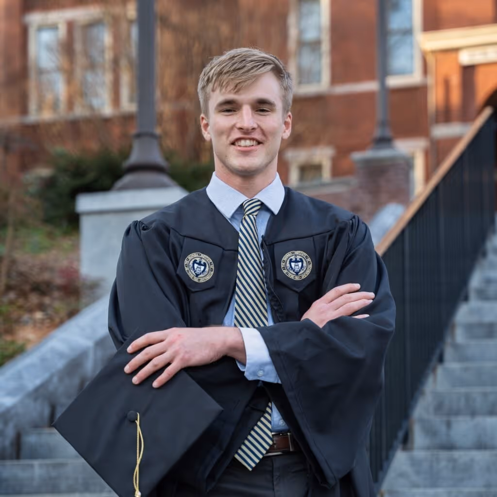 Young man in graduation gown holding a mortarboard, standing on outdoor steps with a brick building in the background.