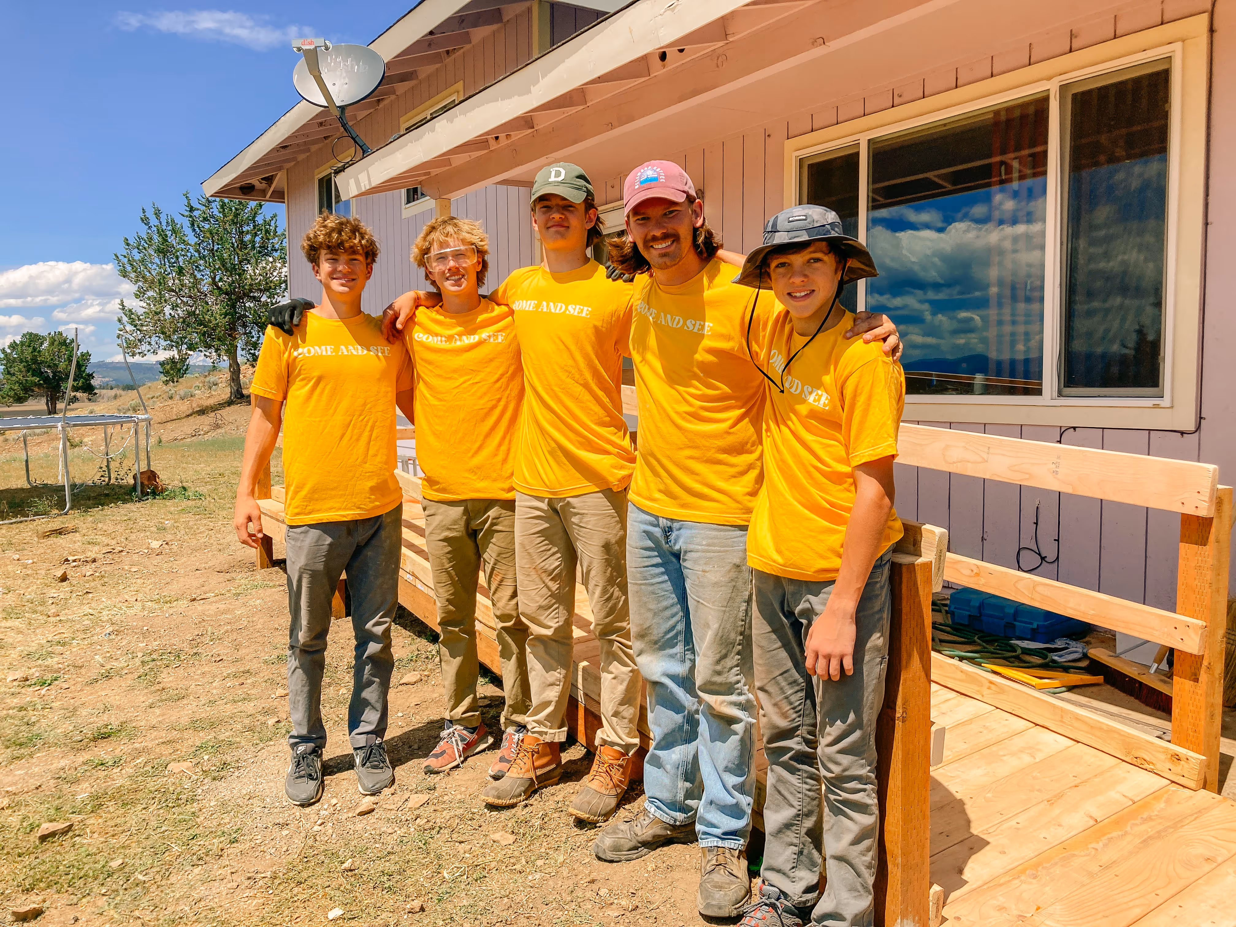 Five young men wearing matching yellow 'COME AND SEE' t-shirts standing arm in arm outside a house on a sunny day.