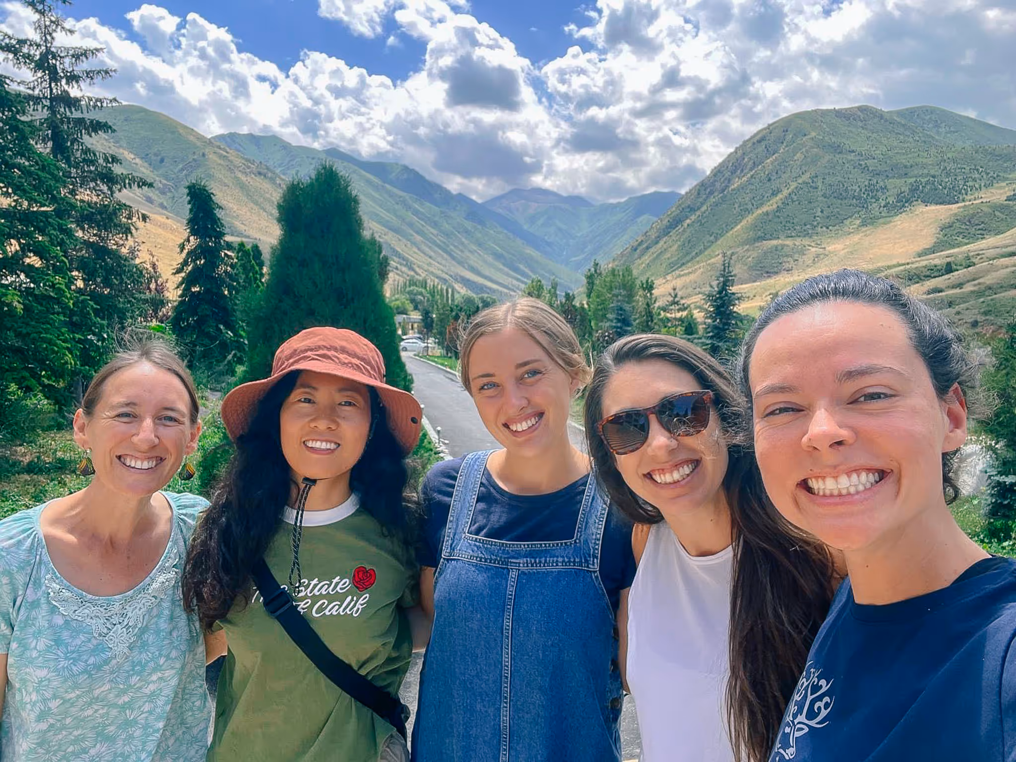 Five smiling women posing for a selfie outdoors with green hills and a partly cloudy sky in the background.