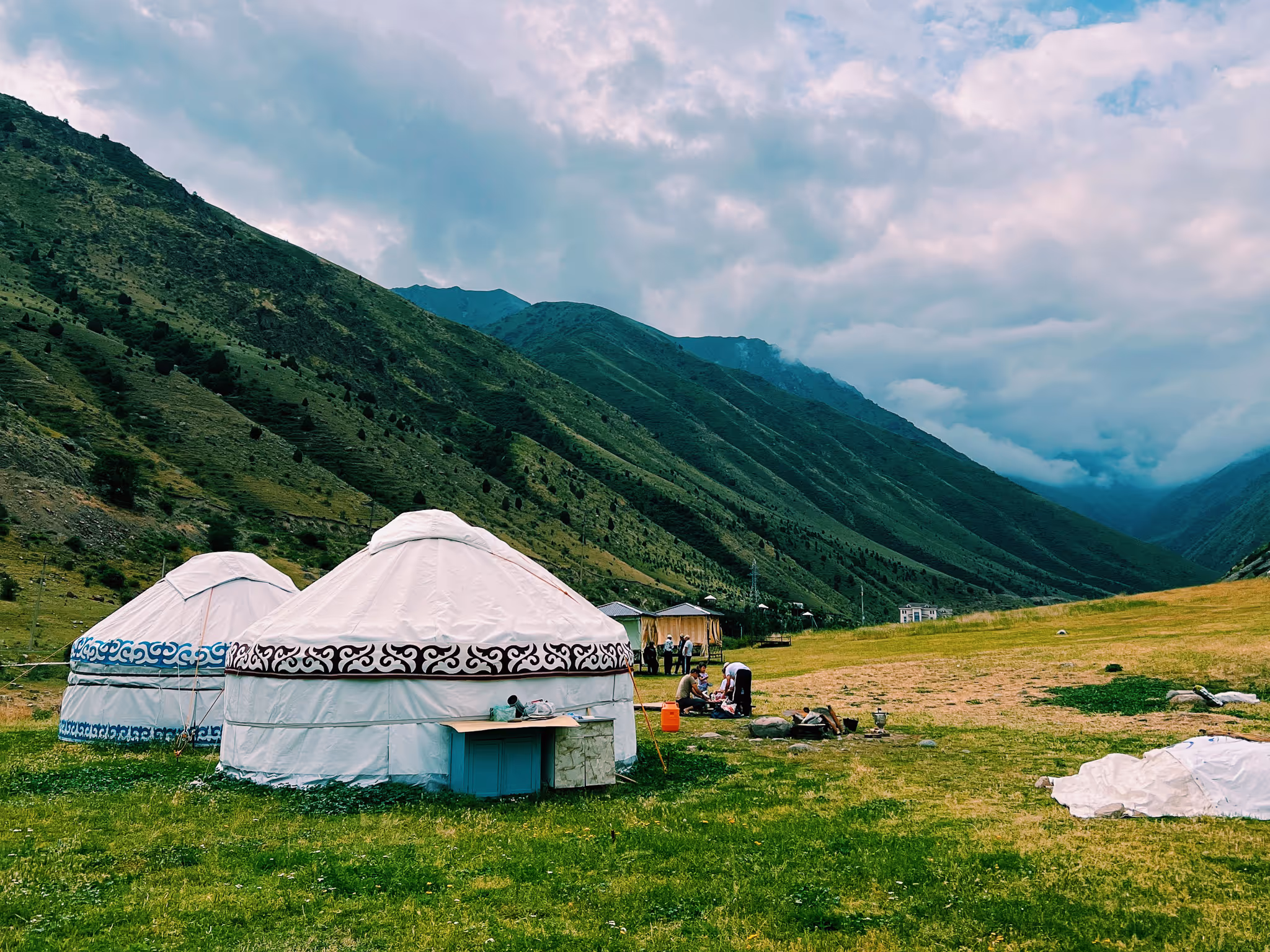 Two traditional yurts with decorative patterns pitched on green grass in a mountain valley under cloudy sky.