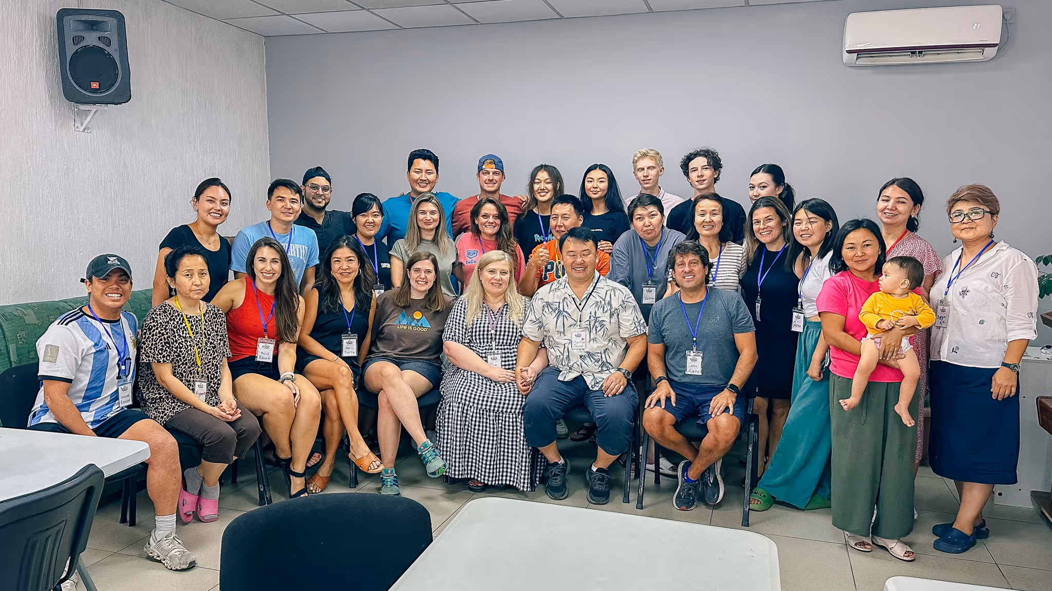 A diverse group of 29 adults and one baby posing indoors, with many wearing name tags and smiling.