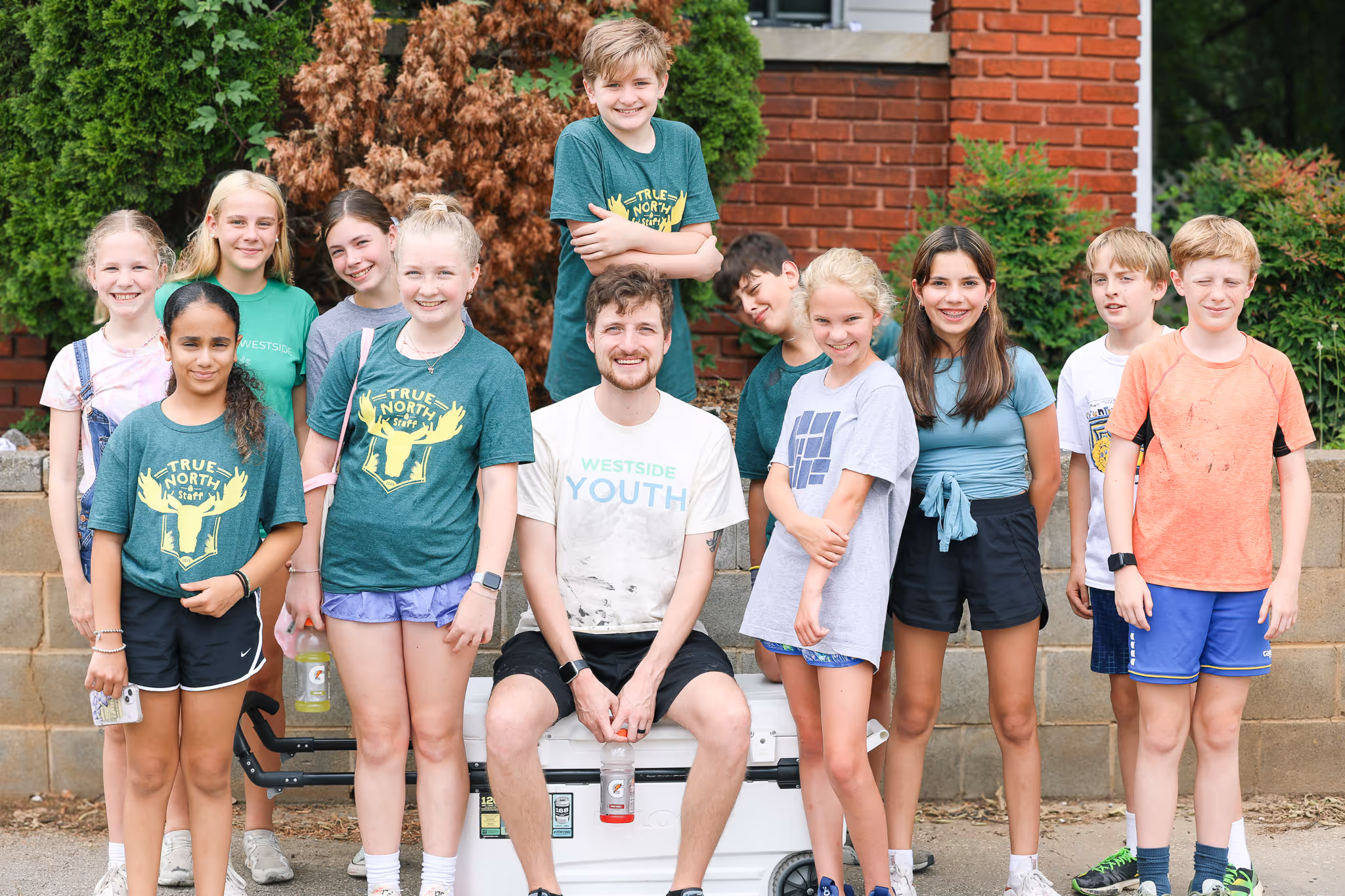 Group of smiling kids and a male youth leader wearing casual sportswear outdoors in front of a brick wall and greenery.