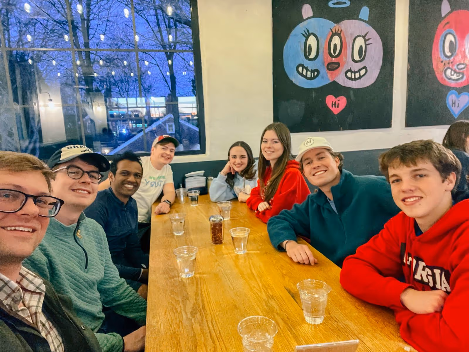 Group of seven young adults sitting closely around a wooden table in a cozy cafe with string lights and colorful cartoon paintings on the wall, smiling at the camera.