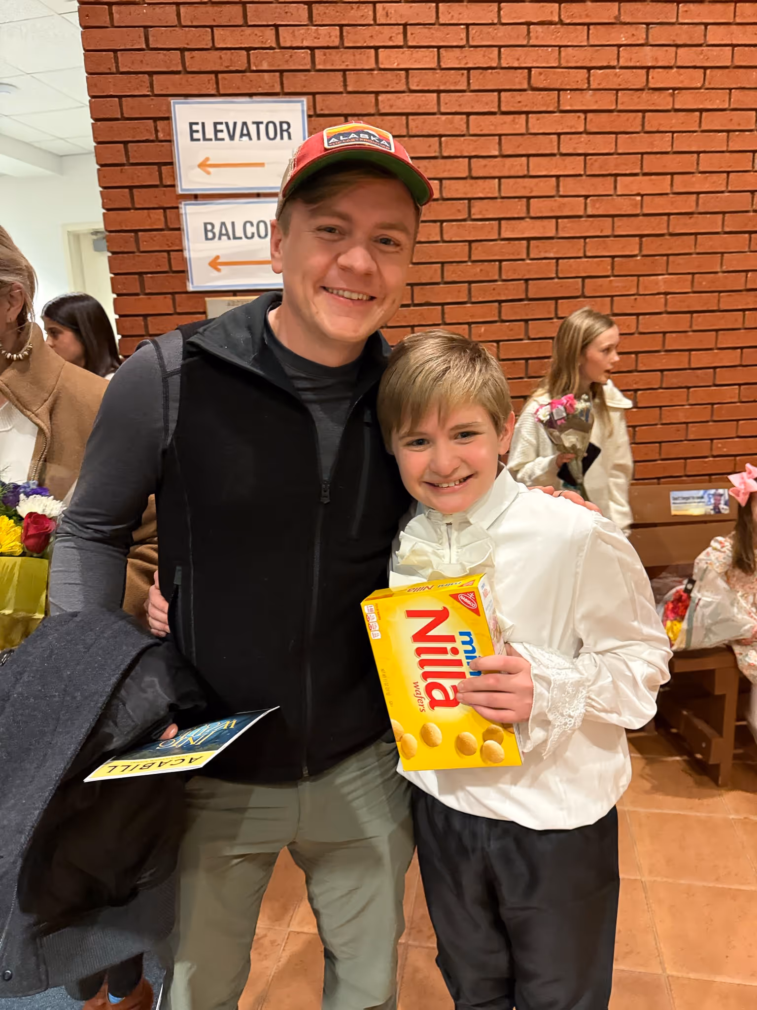 Smiling man wearing an Alaska cap hugs a boy in a white shirt holding a box of Nilla wafers in an indoor setting with a brick wall background.