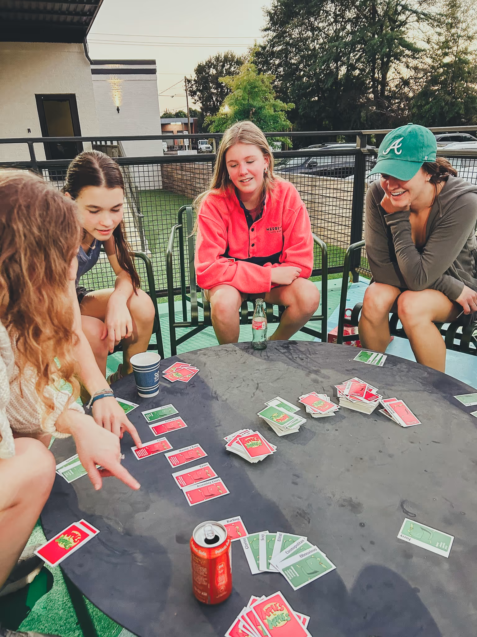 Four young women sitting around a round table playing a card game outdoors on a patio.