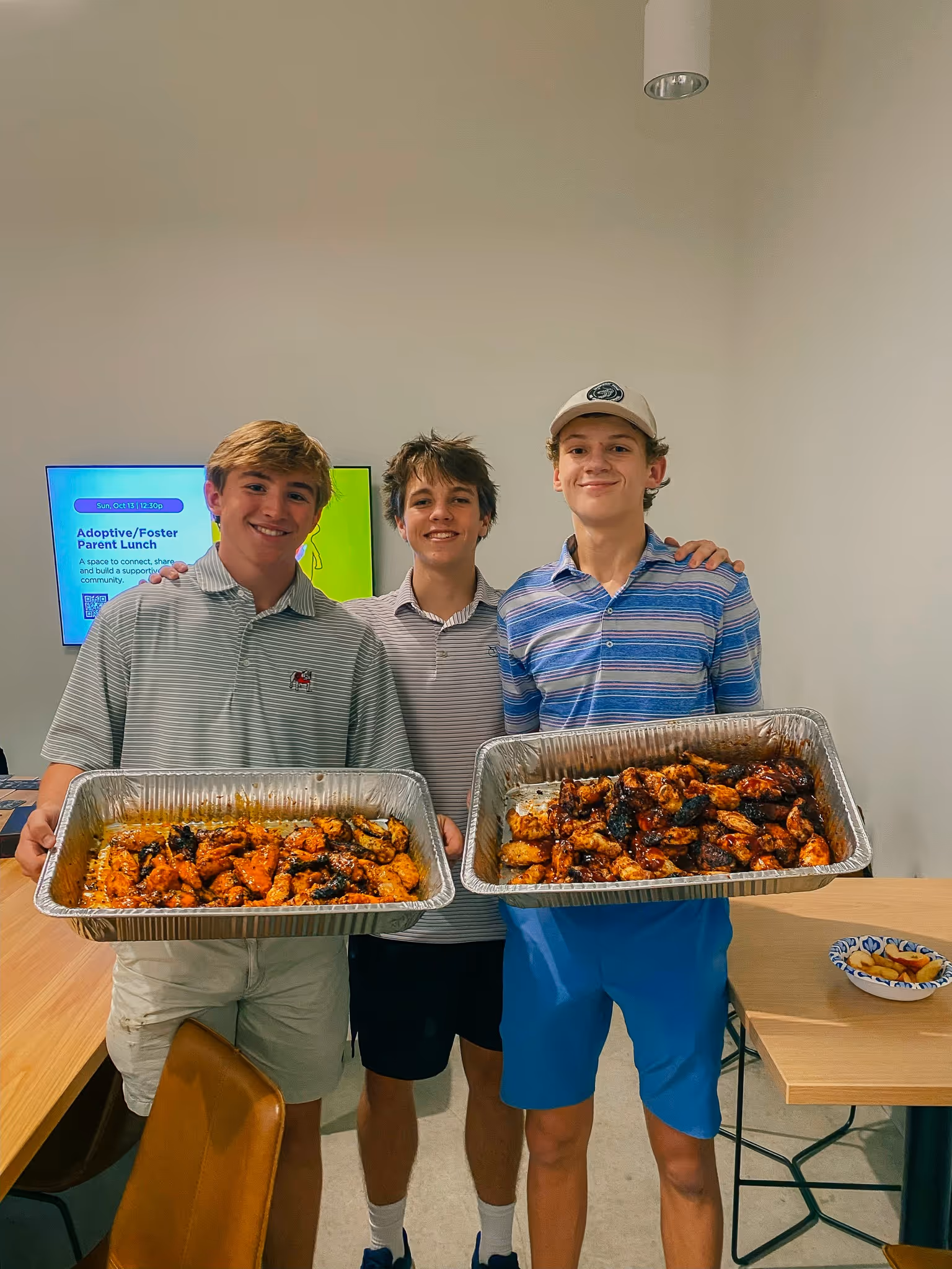 Three teenage boys smiling indoors, two holding large trays of cooked chicken wings.