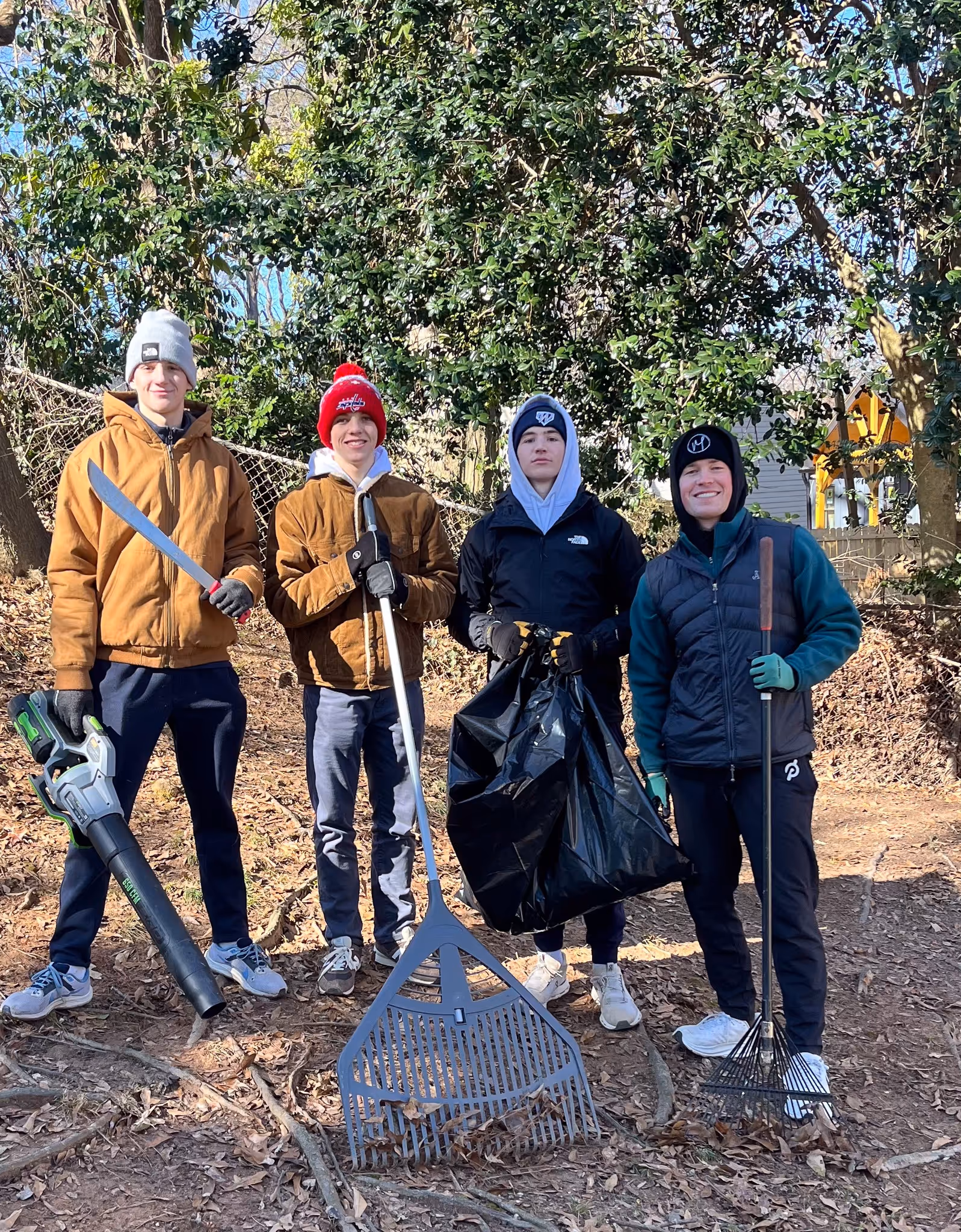 Four young men standing outdoors in a wooded area holding yard tools including a leaf blower, two rakes, and a trash bag, dressed in warm clothing and hats.