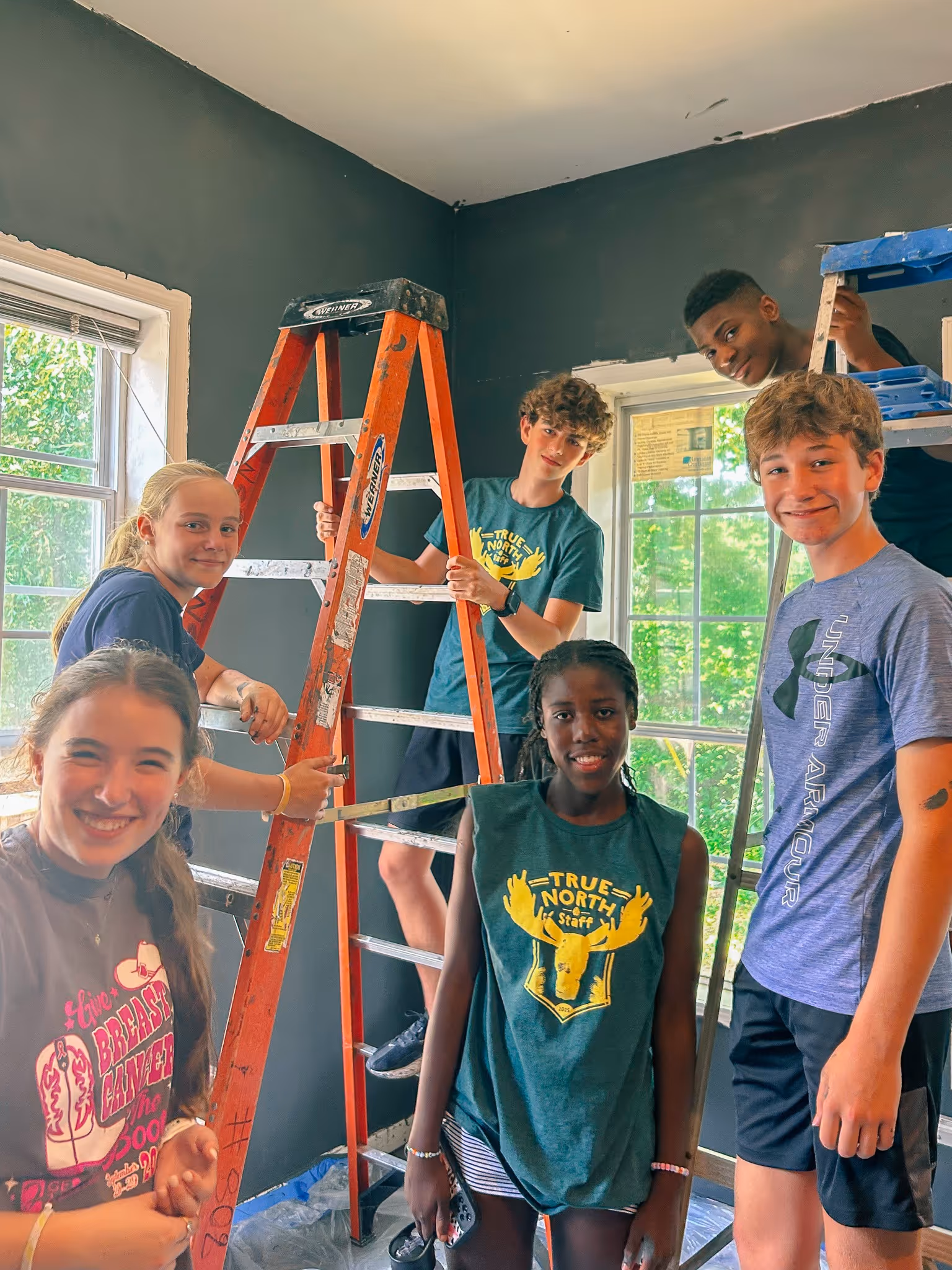 Six smiling teenagers posing inside a room with dark-painted walls, some standing on ladders near windows.