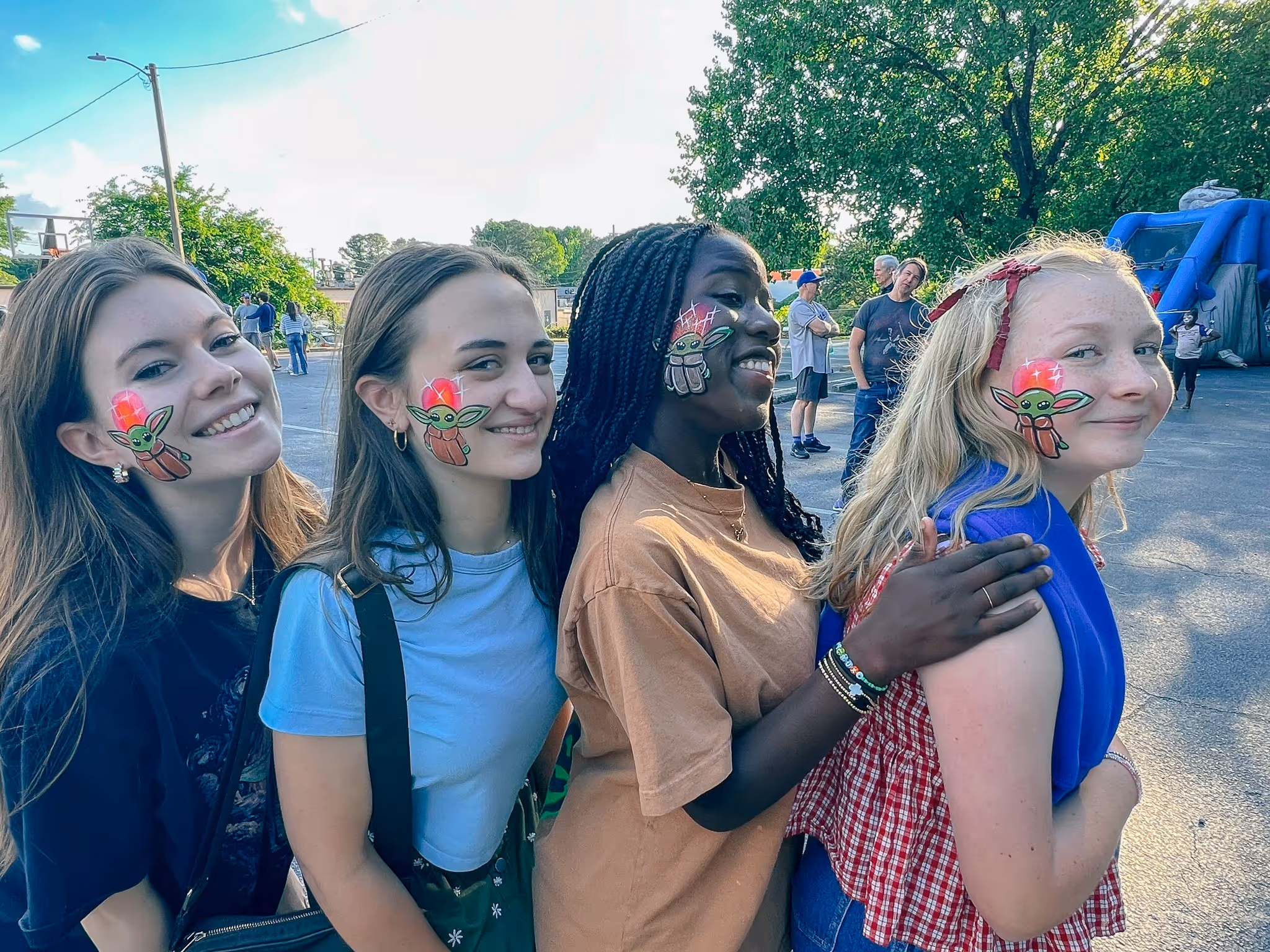 Four smiling young women with Baby Yoda face paint standing closely in a line outdoors at a fair or event.