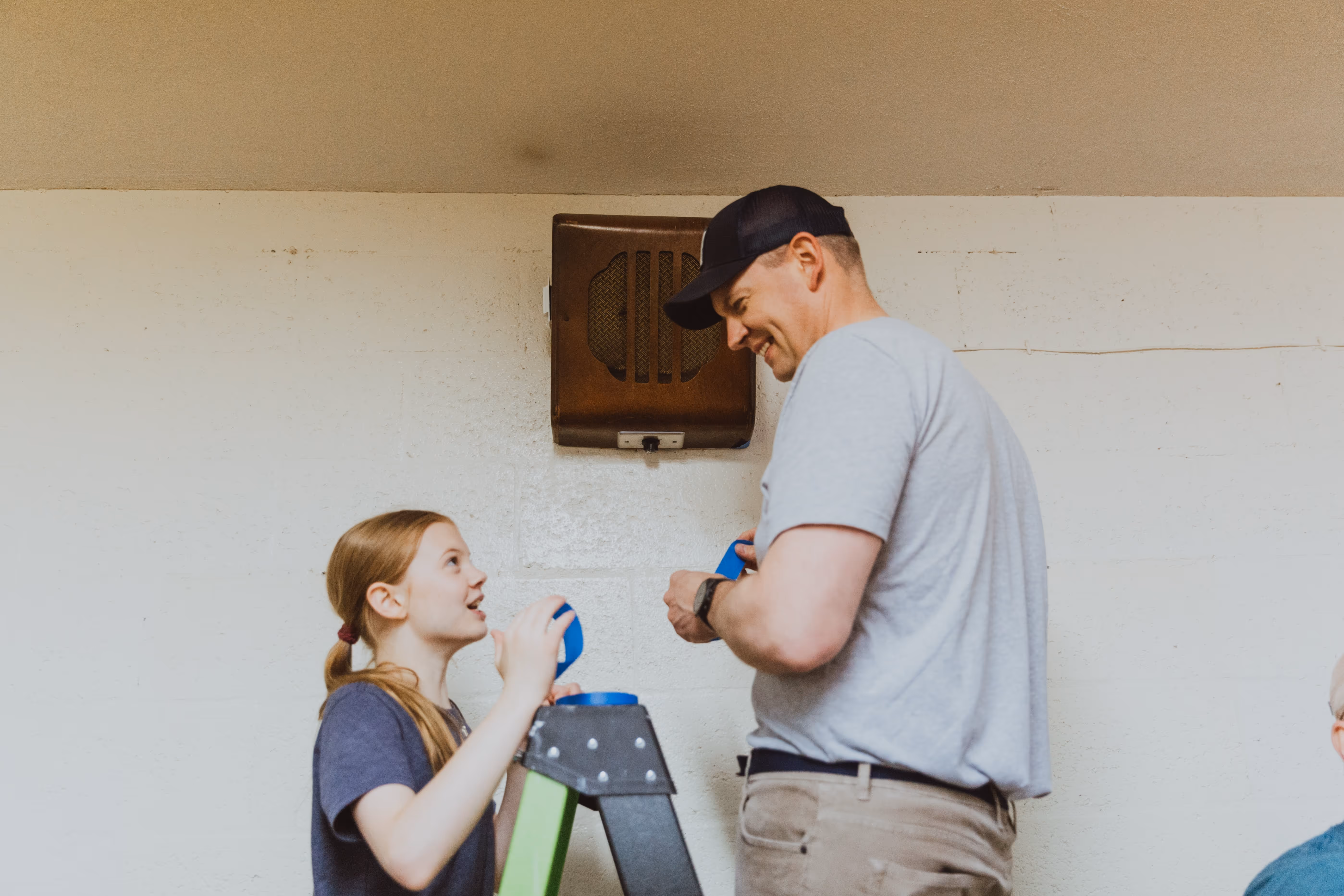 Man and young girl smiling and working together on a home project with a ladder against a white wall.