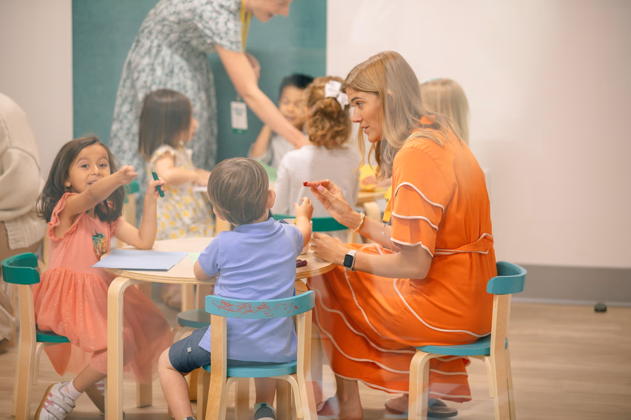 Teacher in an orange dress interacting with young children seated around a small table with drawing materials in a classroom.