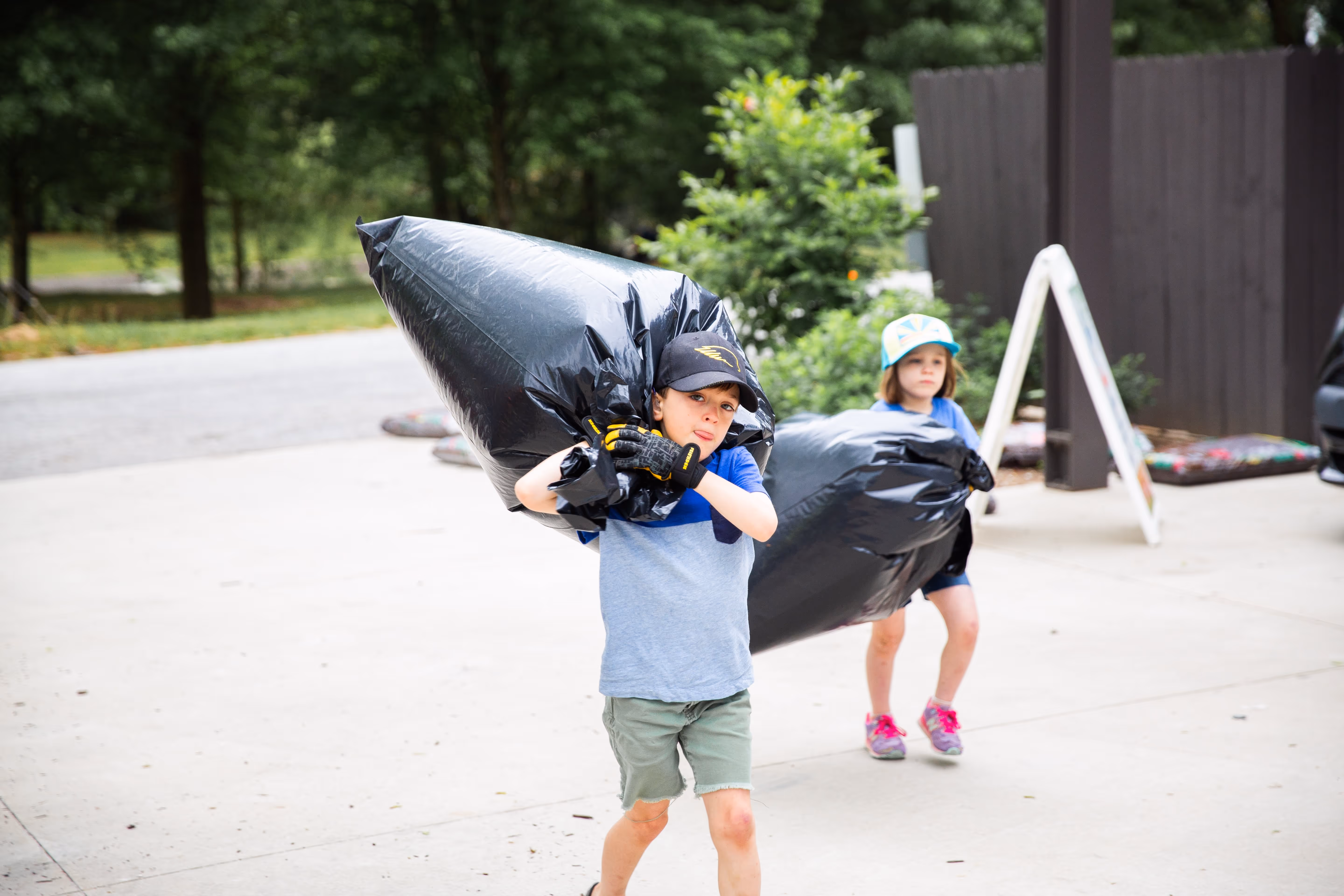 Two children carrying large black trash bags outdoors on a concrete surface.