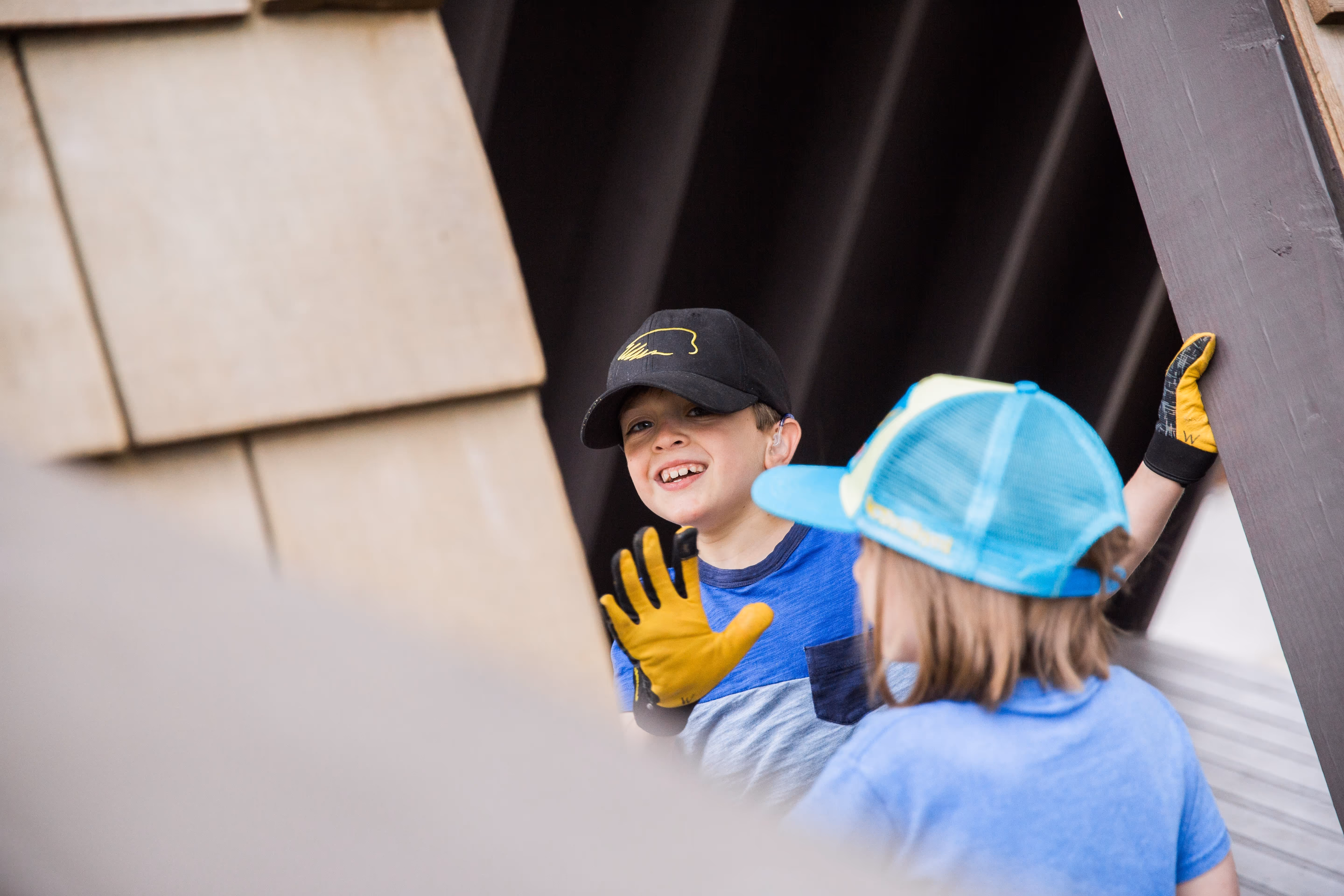 Two children wearing gloves and caps, one smiling and waving inside a wooden structure.