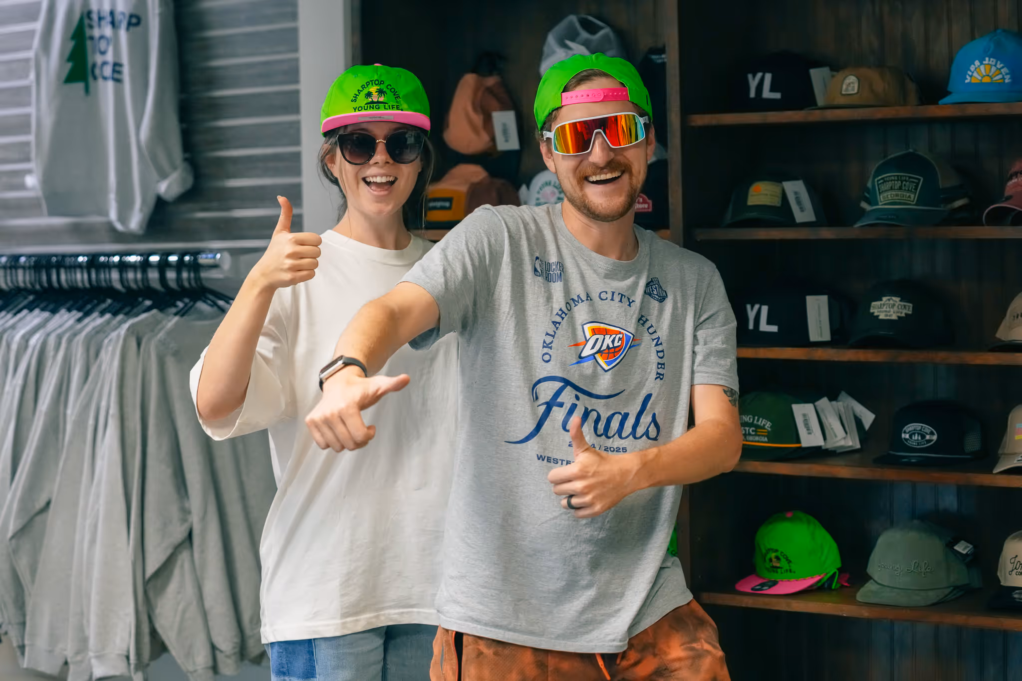 Two people wearing bright green caps and sunglasses smiling and giving thumbs up inside a shop with hats and clothing displayed in the background.