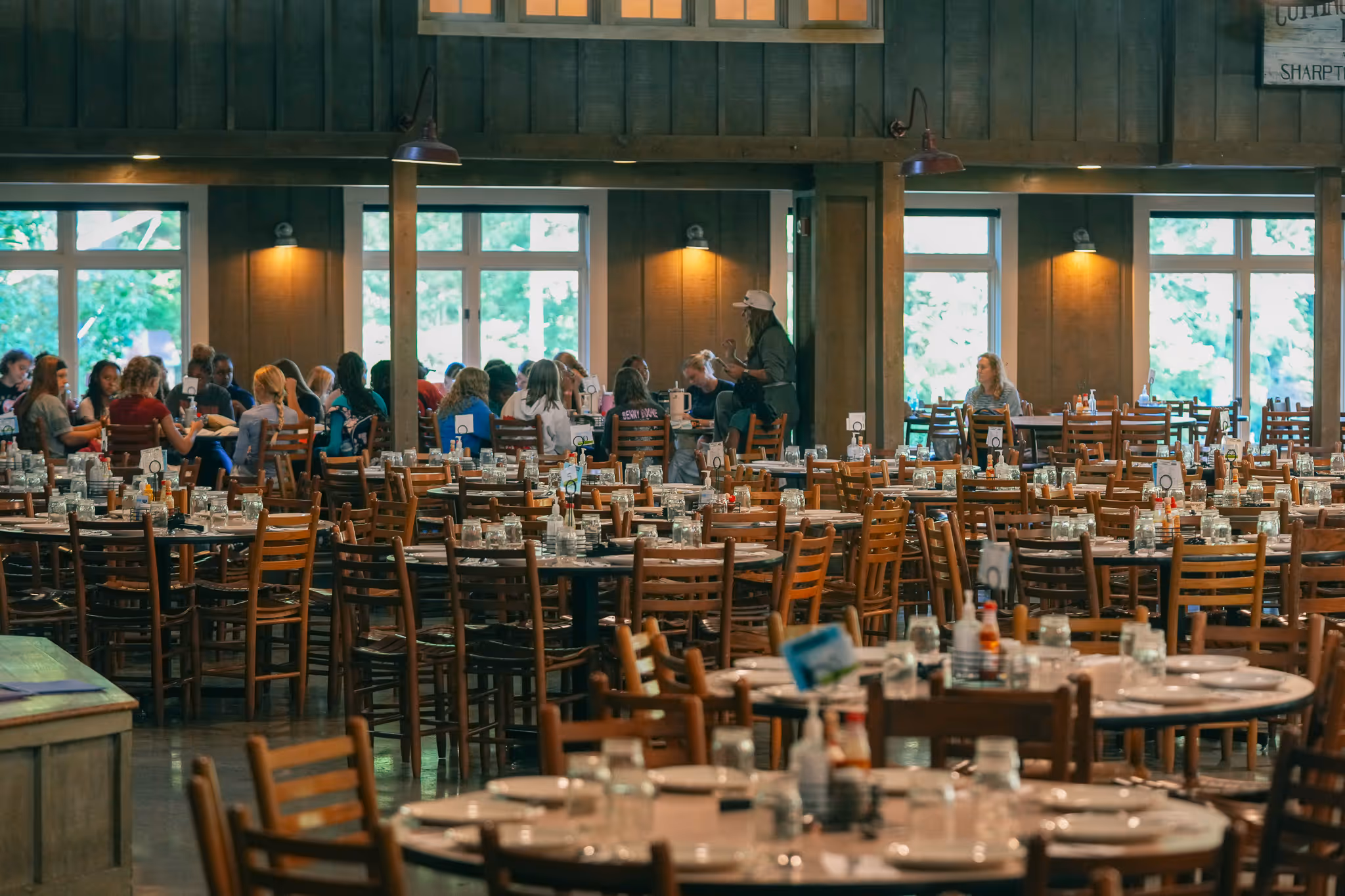Spacious dining hall with many wooden tables and chairs, some occupied by groups of people near large windows.