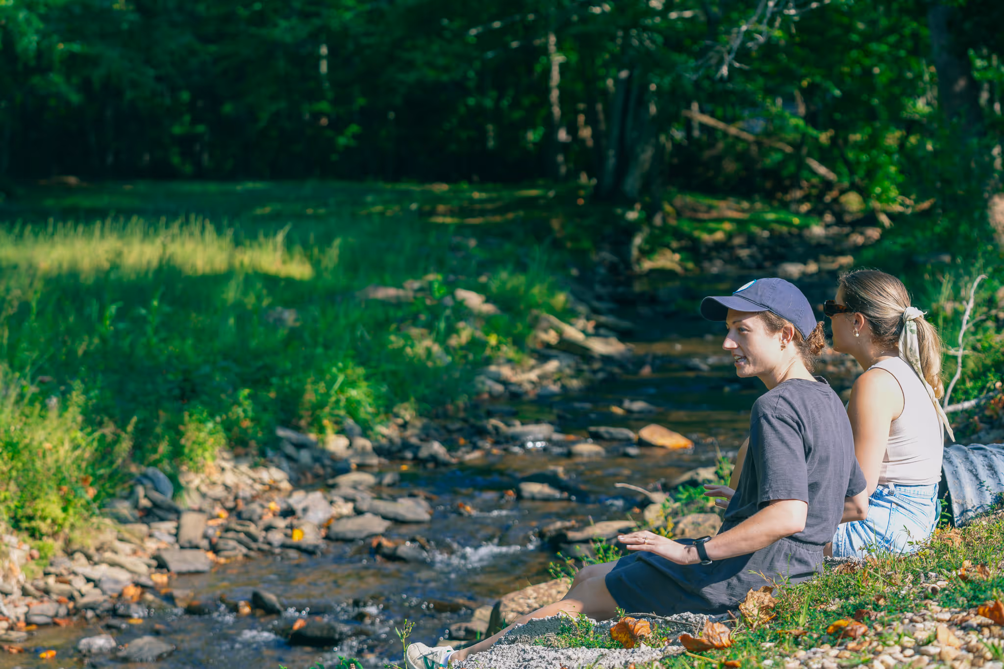 Two young women sitting on a grassy riverbank beside a rocky stream surrounded by green trees.