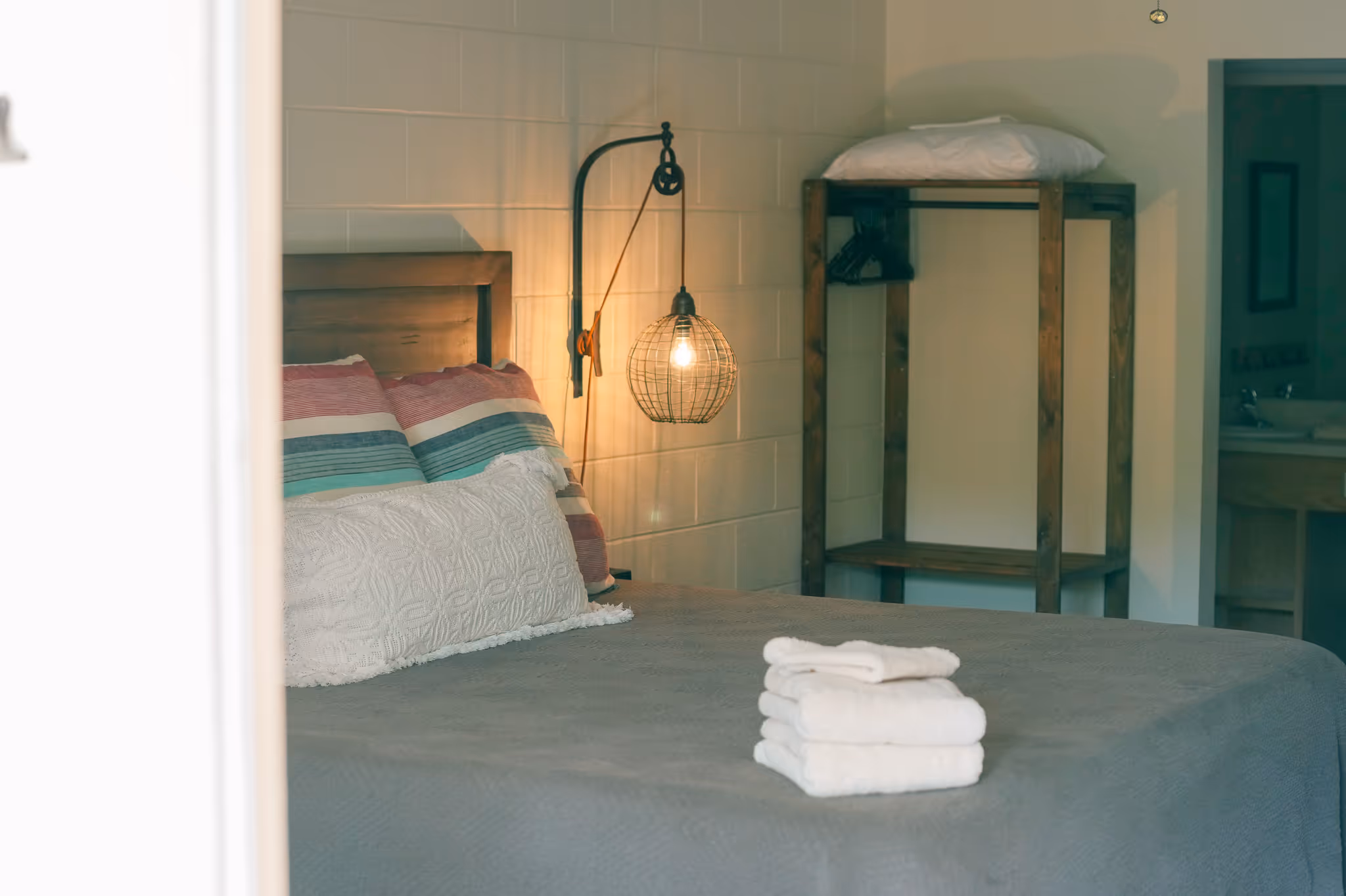 Bedroom with a neatly made bed featuring striped pillows, a quilted white cushion, a stack of folded white towels, a hanging light fixture, and a wooden open wardrobe.