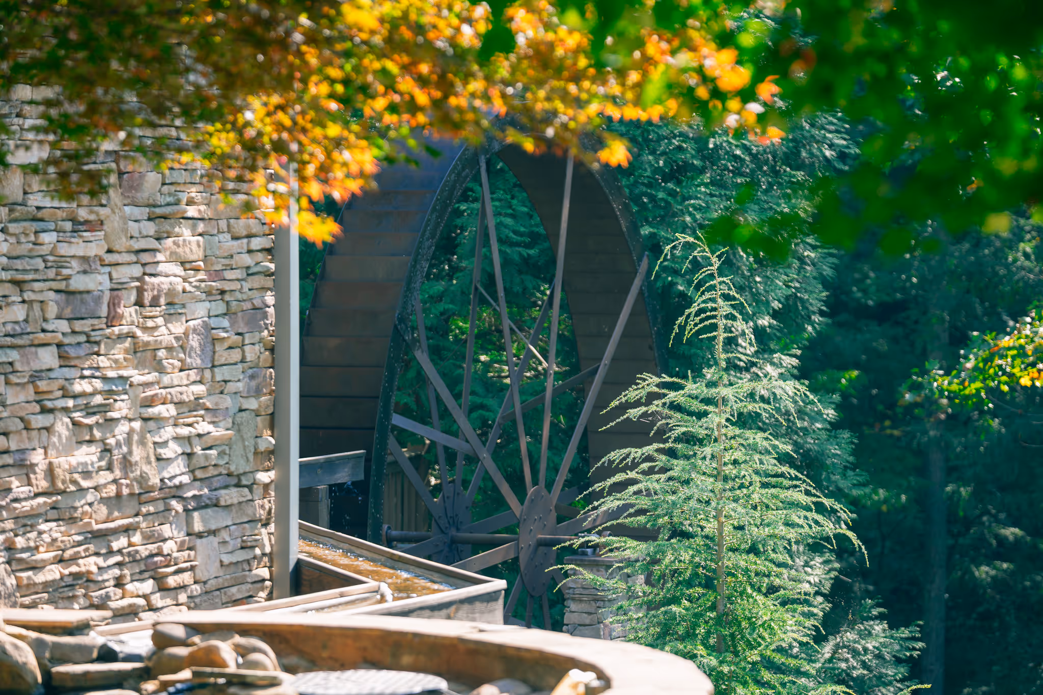 Large metal water wheel attached to a stone building surrounded by green trees and autumn foliage.