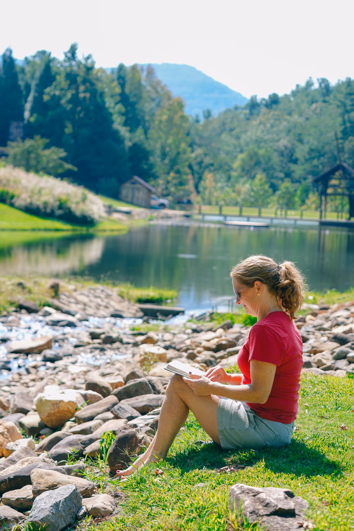 Woman in red shirt and shorts sitting on grass by a rocky pond, reading a book in a sunny park.