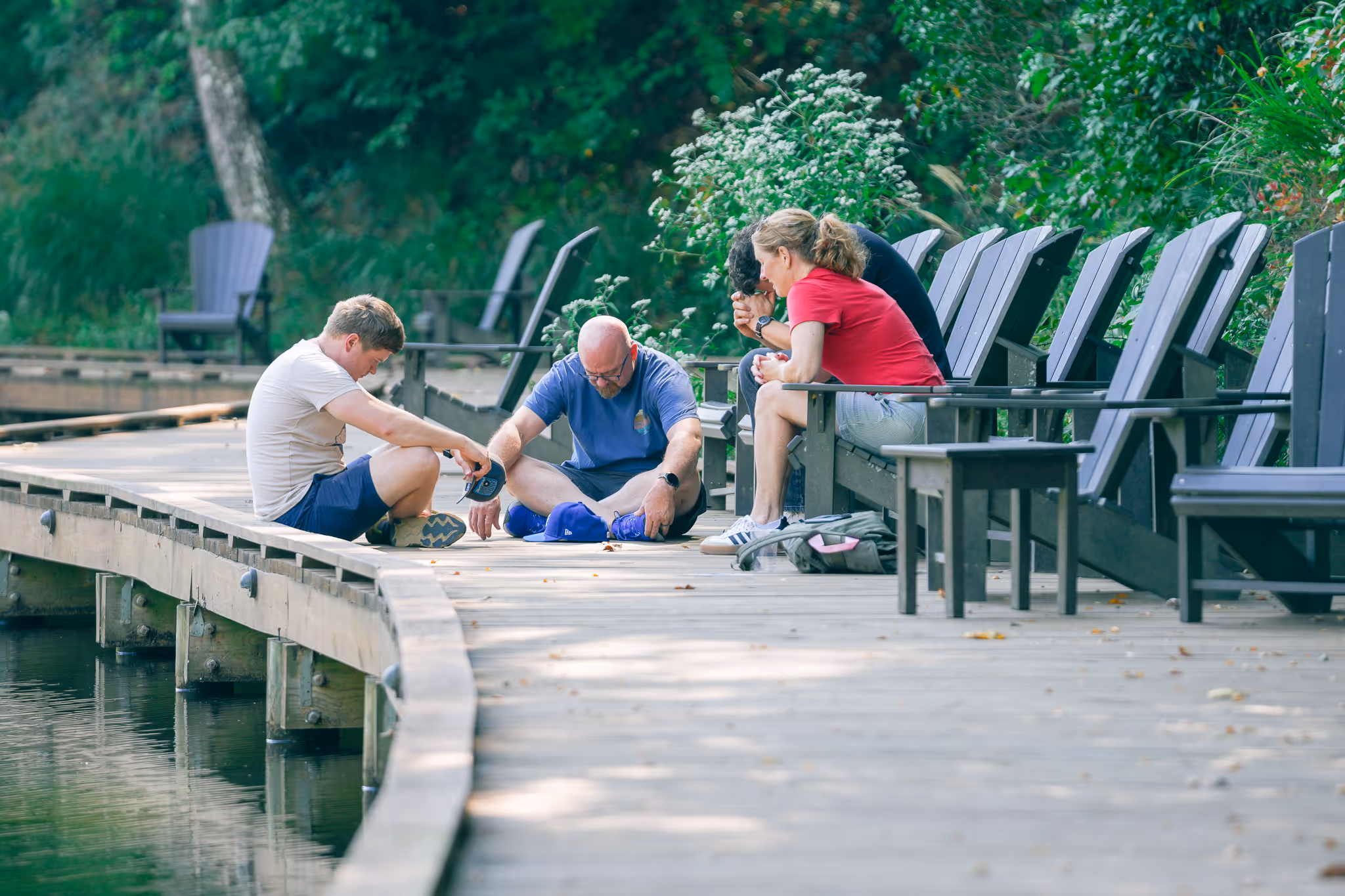 Four people sitting and stretching on a wooden dock beside a lake, with empty chairs and green foliage in the background.