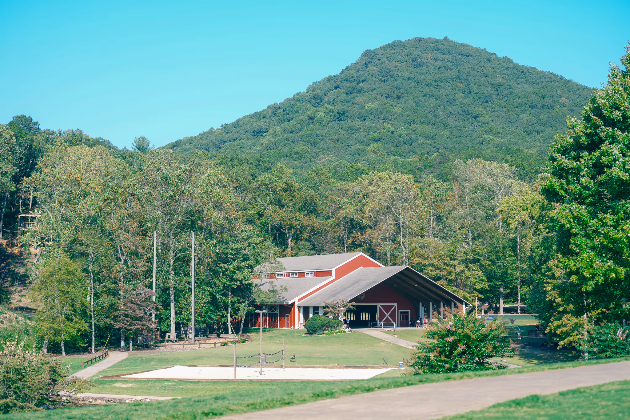 Red barn-style building near a volleyball court with grassy fields and a forested mountain in the background.